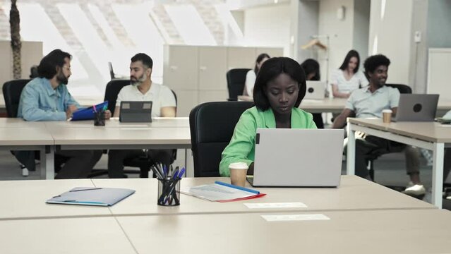 Focused young African businesswoman working at her desk on laptop in modern office with colleagues 