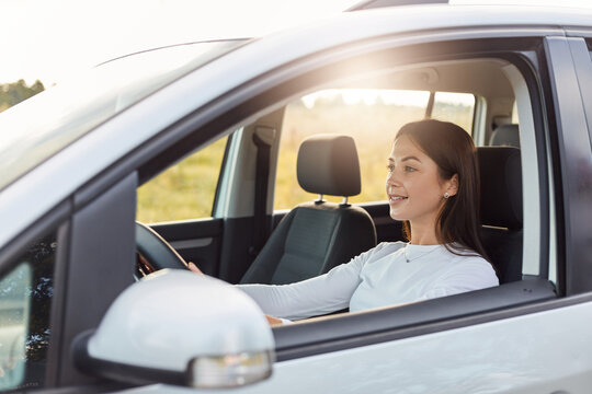 Image of beautiful dark haired woman with happy expression driving car, wearing white shirt, looking at road with charming smile, enjoying, like traveling,