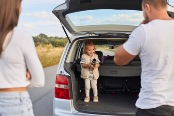 Portrait of happy toddler baby girl standing car trunk and holding thermos, mother and father...