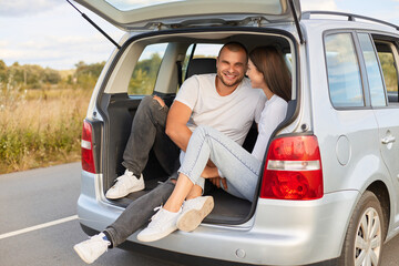 Portrait of loving couple sitting in car trunk wearing white shirts traveling together, talking and smiling, being fall in love, enjoying spending time together.