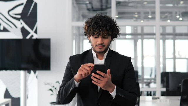 Confident man using mobile phone app in modern office. Indian business professional browsing financial news, smiling reads good news. Successful male staying up to date with latest news and trends. 