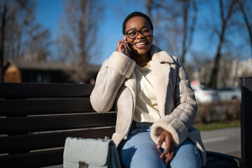 Outdoor portrait of happy african-american woman on sunny day. She is sitting on bench in the street and talking on phone.
