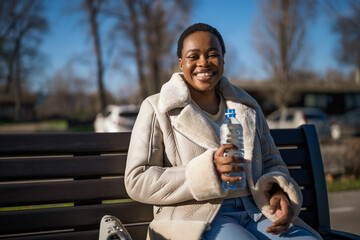 Fototapeta premium Outdoor portrait of happy african-american woman on sunny day. She is sitting on bench in the street and drinking water.