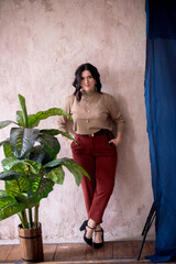 Young stylist girl with dark hair smiling in mocha golf trendy gold jewelry and brown trousers in a light studio with green live plants in pots