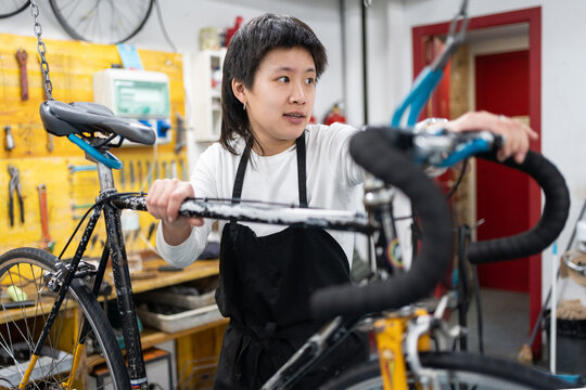 portrait of smiling asian girl behind hanging bicycle, young woman fixing a bike in a collective self-repair shop. girl in black non-binary apron