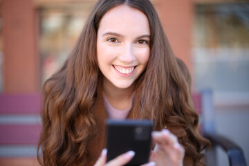 Happy woman on a bench looks at you holding phone