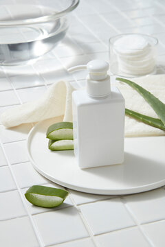 A pump bottle dispenser decorated with Aloe vera slices on a round dish, a glass bowl of water and cotton pads displayed. It helps in gentle cleansing, and its antimicrobial properties treat pimples