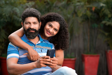Happy indian couple sitting at out of home and using smartphone together.