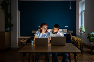 Fototapeta na wymiar Young diverse entrepreneurial couple running small online business at home, multiracial family man and woman sitting at table working on laptops together in evening. Family entrepreneurship concept