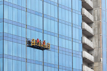 Construction workers on a suspended cradle mount the glass facade of a multi-story skyscraper. - 588255597