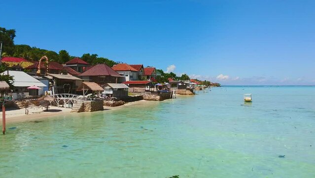 Idyllic ocean scenery by beach with traditional houses and palm trees. View over blue sea and island in summer. Nusa Lembongan, Selat Ceningan, Bali, Ocean side houses, beach. Drone flying forward. 4K