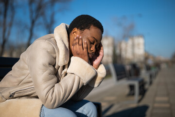Outdoor portrait of depressed african-american woman on sunny day. She is sitting on bench in the street.