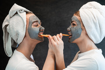 two caucasian women in white towel applying mud facial mask and hold orange isolated on black - obrazy, fototapety, plakaty