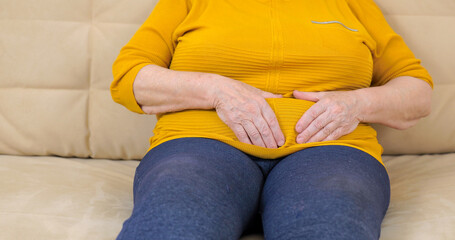 Elderly fat woman having painful stomach ache sitting on sofa.