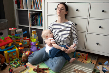 tired sad mother with a small baby todler in her arms sits in a mountain of toys in a mess and...