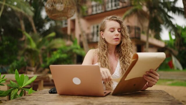Caucasian woman with laptop sitting in outdoor cafe at resort. Female freelancer looking at menu at tropical location. Young adult working remotely at exotic location checks the menu choosing food