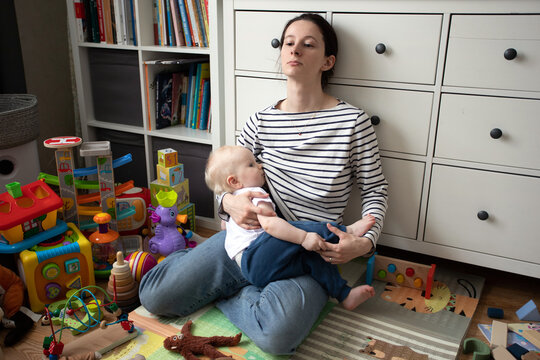 tired sad mother with a small baby todler in her arms sits in a mountain of toys in a mess and breastfeeds