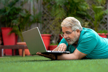 Indian senior man laying down at ground and using laptop.