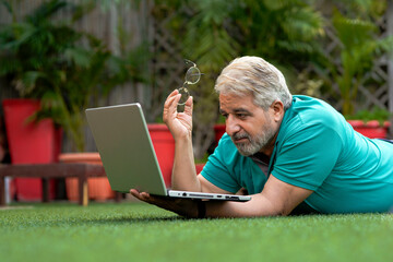 Indian senior man laying down at ground and using laptop.