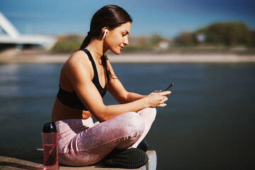 Fototapeta na wymiar Young fitness woman taking a break after jogging and listening to music on smartphone