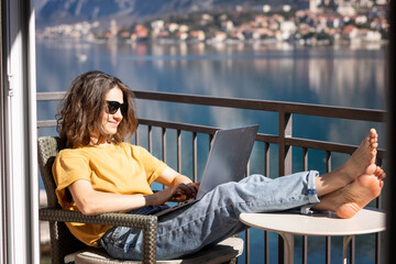 Beautiful young girl woman in sunglasses sitting with a laptop on her balcony with a view of the city, remote work from home, a successful freelancer.