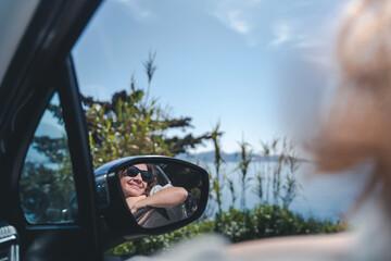 Happy cheerful young woman traveling on the sea by car. Portrait in the reflection of the rearview mirror. Summer vacation concept