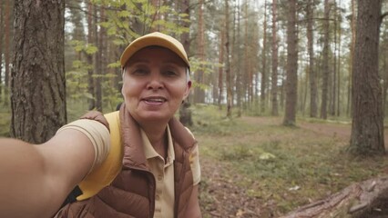 Handheld POV shot of cheerful active senior Asian woman recording video blog while hiking in forest in late summer