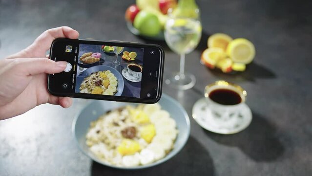 A female blogger takes pictures of breakfast on her phone. Healthy morning food with fruit, porridge, muesli, donuts and coffee at home. High quality 4k footage