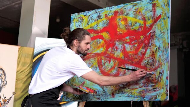 Expressive artist, wearing a white t-shirt and black apron, is creating a masterpiece on canvas in studio. Man applying blue paint ontoabstract painting, demonstrating artistic skills and techniques.
