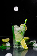 Falling Ice Cubes into a glass of Mojito on a black background. Refreshing Mojito Cocktail with lime, ice cubes and green mint leaves