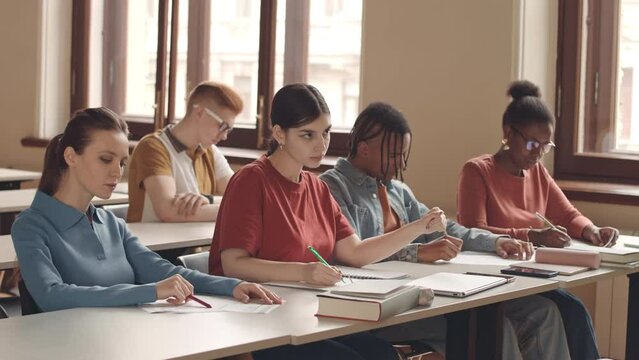 Waist up slowmo of multicultural group of students having lecture in auditorium. Biracial girl raising her hand to ask professor question