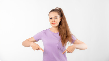 Happy young woman with long hair pointing fingers down and smiling, showing advertisement, product below, promo offer, standing in casual t shirt over white background