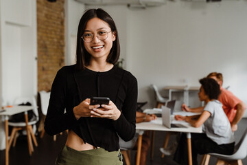 Cheerful asian woman using smartphone while standing in office with her colleagues on a background