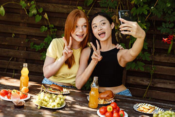 Two beautiful women taking selfie and showing peace sign during barbeque at backyard