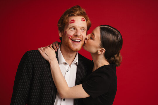 Woman kissing man with red lipstick marks on his face isolated over red wall