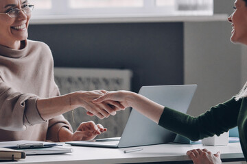 Two confident women shaking hands and smiling while sitting at the office desk together