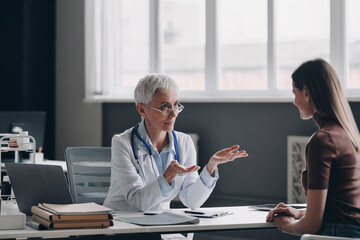 Obraz premium Senior female doctor talking to patient and gesturing while sitting at the medical office
