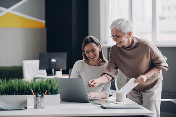 Two confident businesswomen looking at laptop and smiling while working in office together