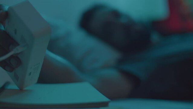 Young man lying in bed in dark room, staring up and checking time on clock while suffering from insomnia