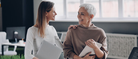 Two happy businesswomen communicating and smiling while standing in the office together