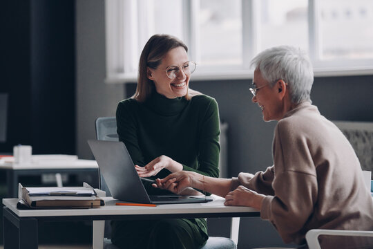 Happy financial advisor pointing laptop while discussing options with senior woman in office