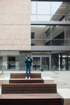 Businesswoman talking on smart phone standing on bench outside building