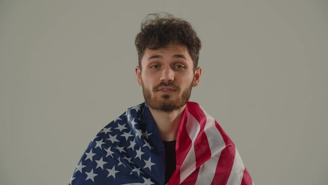 A young brunette standing with an American flag looks questioningly into the camera. Close-up of a young male activist with an American flag posing against a gray background.