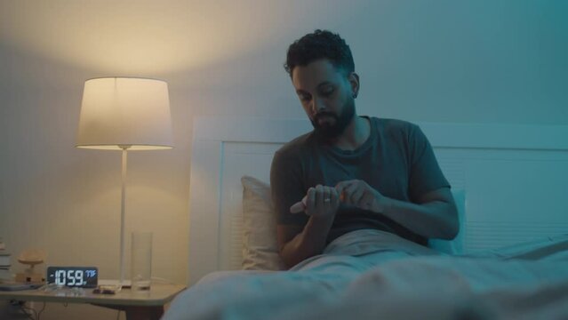 Medium shot of man taking sedative pills, drinking water, then turning off lamp and lying under blanket in bed while going to sleep at night