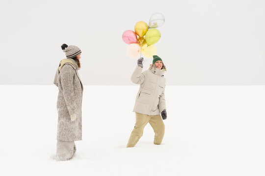 Smiling woman with shiny balloons having fun with friend in snow