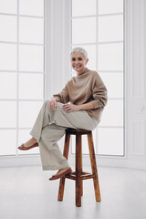 Confident senior woman looking at camera and smiling while sitting on the chair against a window
