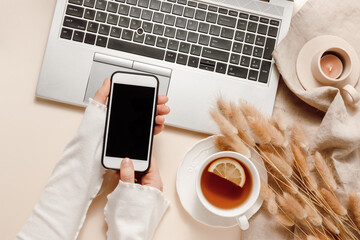 Woman hands hold mobile phone at laptop background with tea, candle, lagurus grass on linen cloth, aesthetic home workplace, cozy feminine office desk. Online shopping, work and business branding