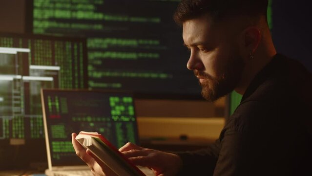 Close-up view of a young man sitting in his room in front of several monitors and reading a book. A young man reads literature for beginner programmers.