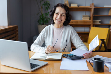 Fototapeta na wymiar Middle aged senior woman sit with laptop and paper document. Smiling older mature lady reading paper bill pay online at home managing bank finances calculating taxes planning loan debt pension payment
