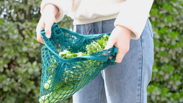 Female hand holding reusable shopping bag with vegetables on green background, zero waste concept.